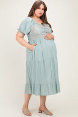woman wearing a light blue maternity dress with floral embroidery and pockets, gently cradling her baby bump.