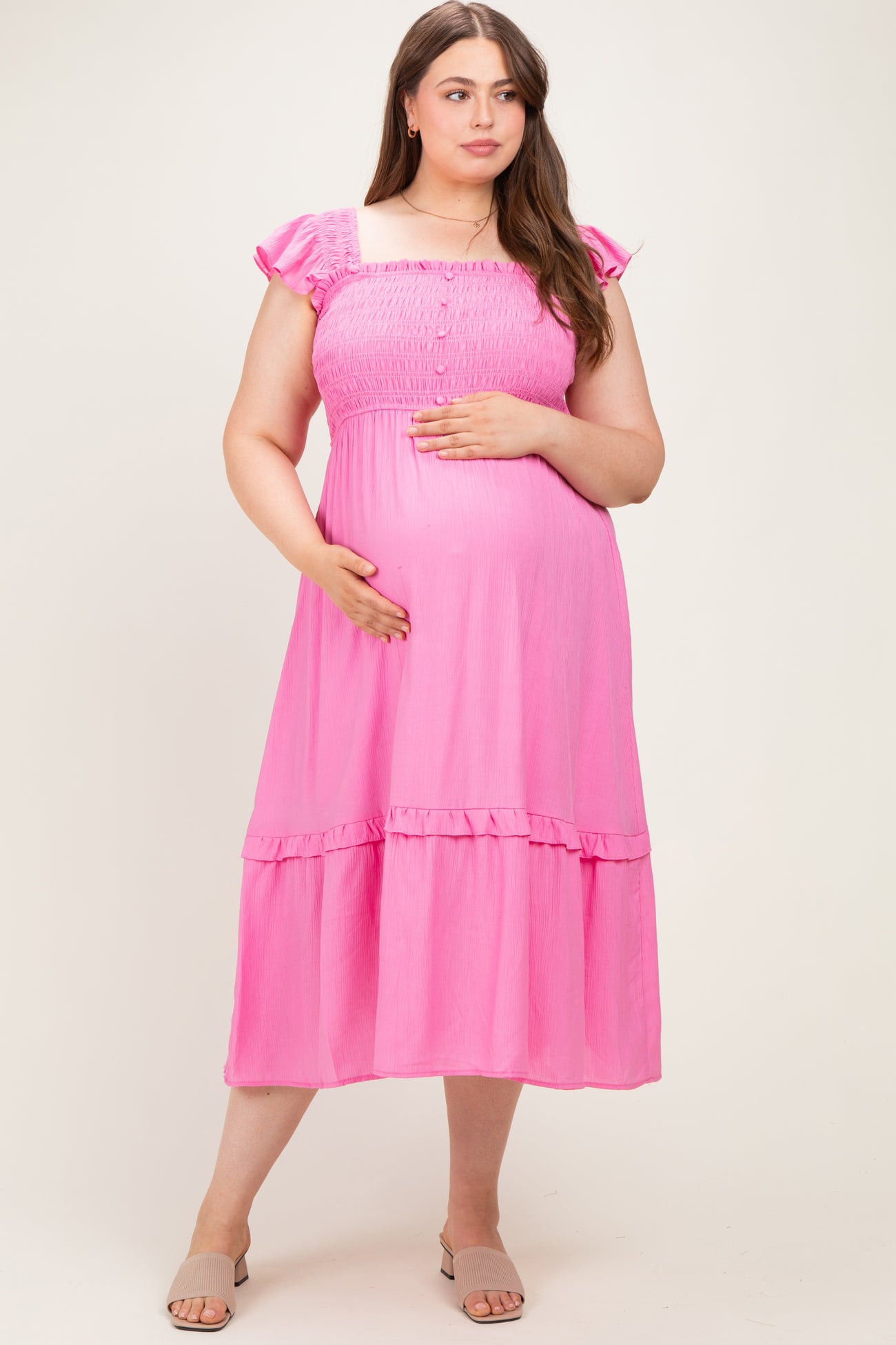 woman wearing a pink maternity plus dress with smocked bodice and ruffled sleeves, holding her baby bump