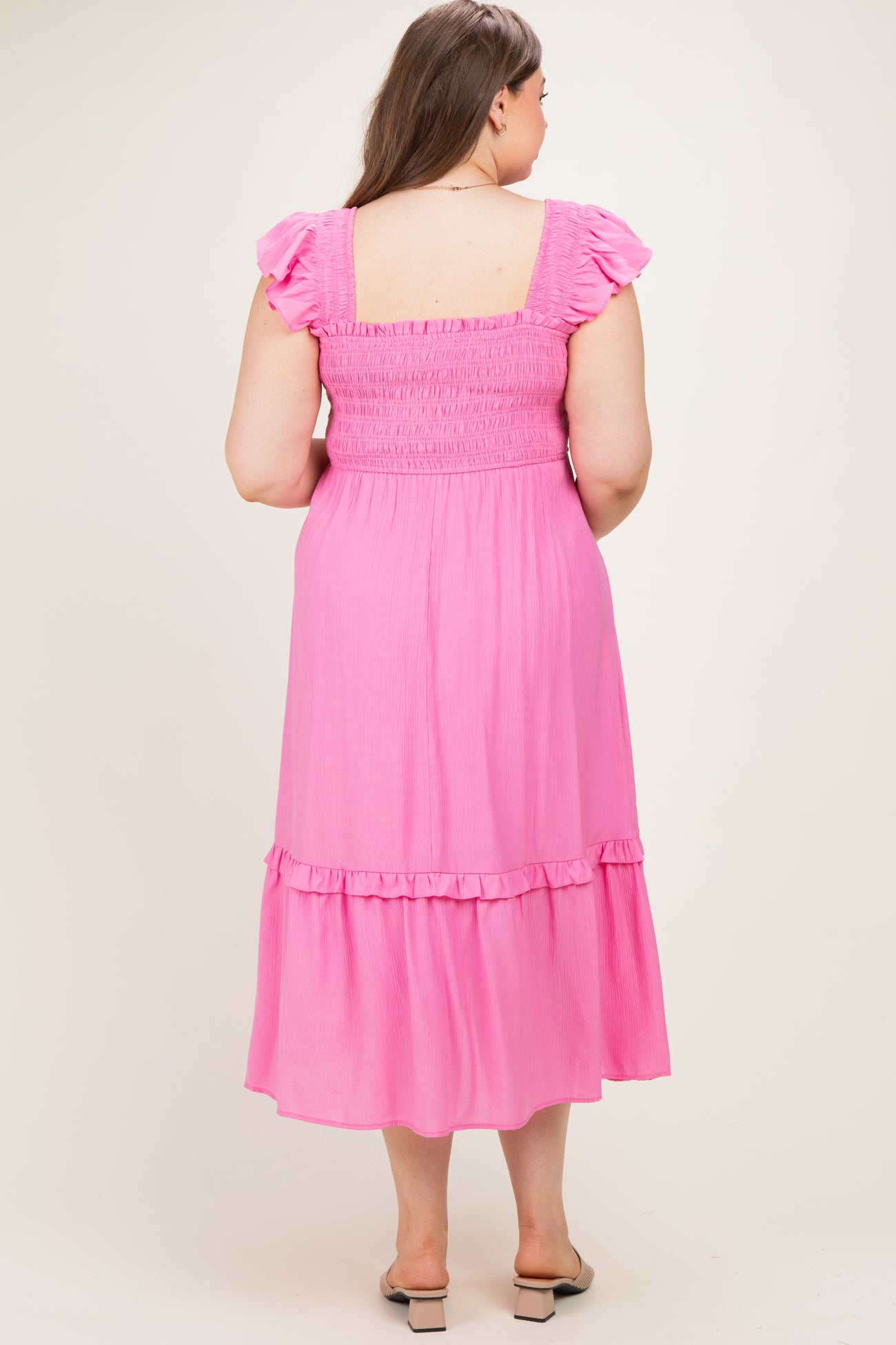 woman wearing a pink maternity plus dress with ruffle sleeves and a tiered skirt, showing her baby bump from the back