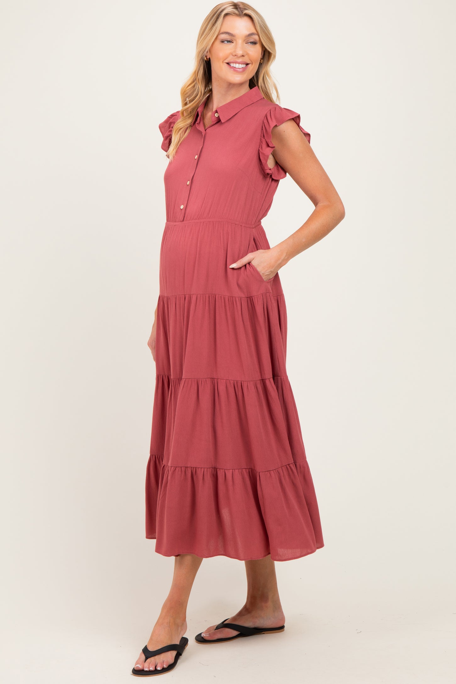 woman wearing a coral pink maternity dress with ruffled sleeves and a tiered skirt, hands in pockets and smiling