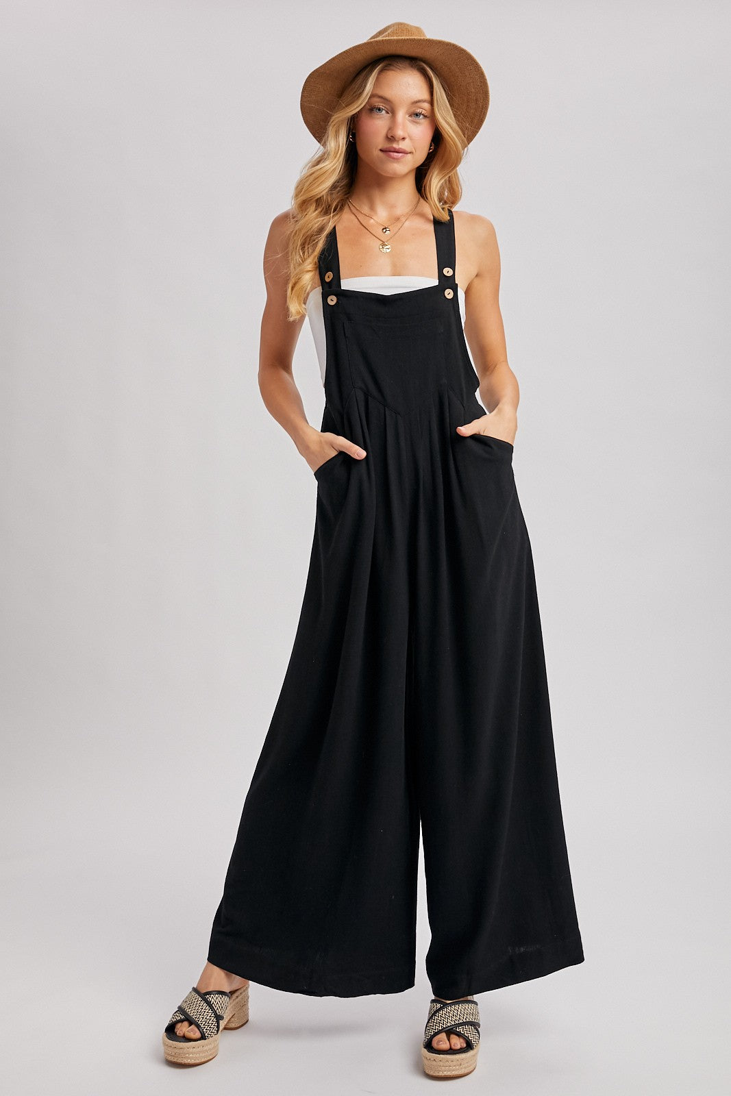 woman wearing a black wide-leg jumpsuit with shoulder straps and pockets, styled with a white tank top and a straw hat