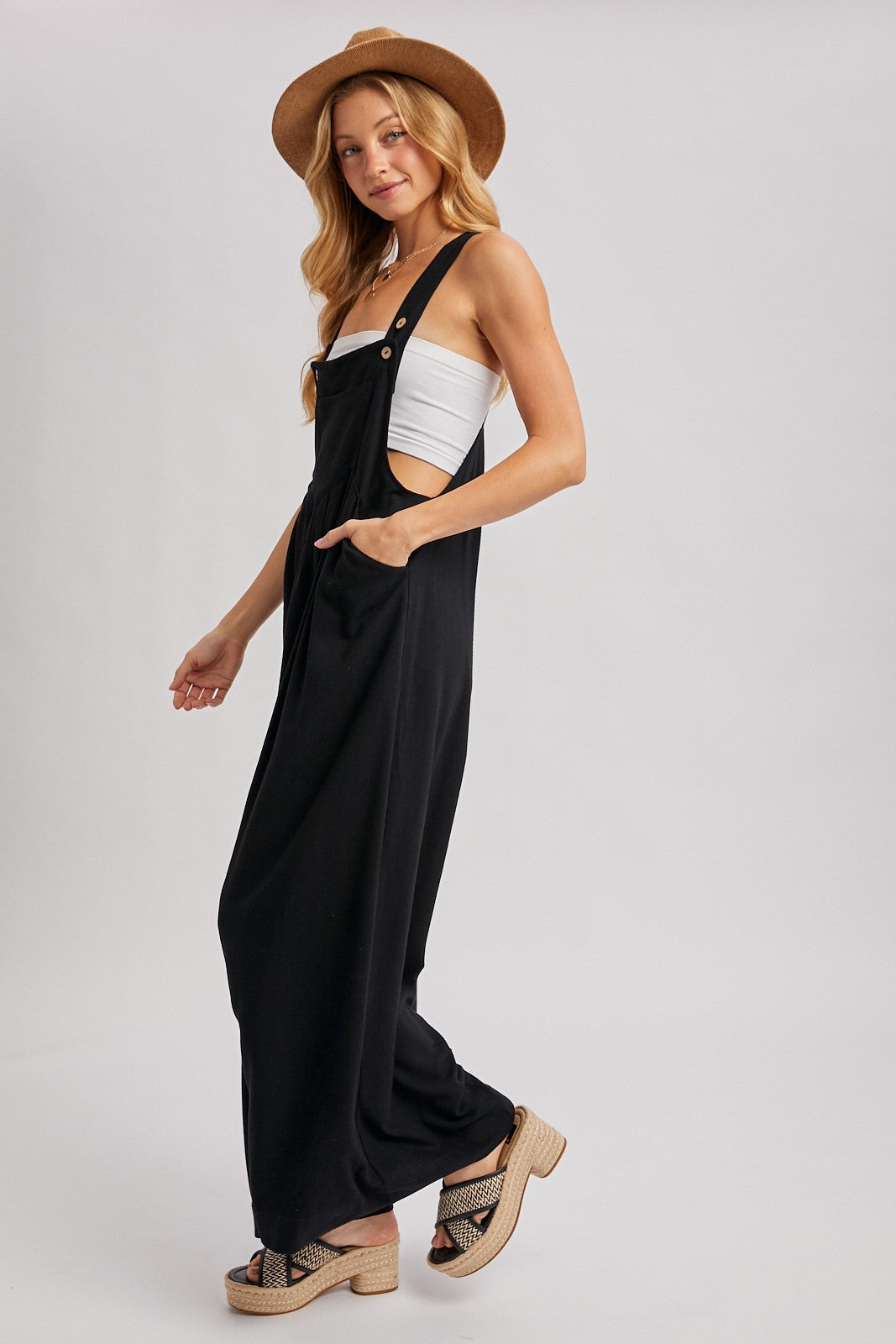 woman wearing a black jumpsuit with side cutouts, buttons, and pockets, paired with a white top and a straw hat