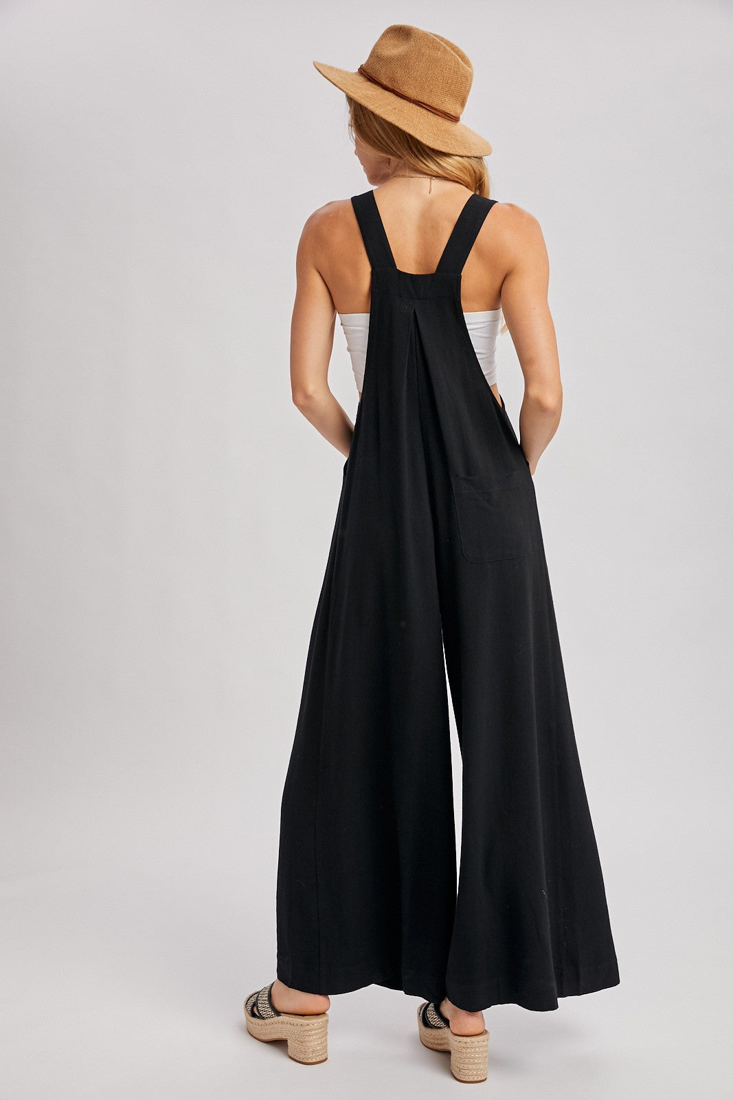 woman wearing a black wide-leg jumpsuit with adjustable straps and a pocket, facing away and wearing a straw hat