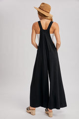 woman wearing a black wide-leg jumpsuit with adjustable straps and a pocket, facing away and wearing a straw hat