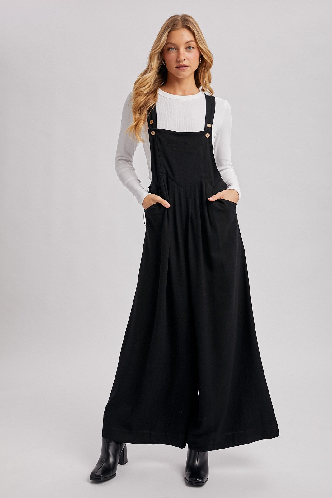 woman wearing a black wide-leg overall dress with pockets and gold button details over a white long sleeve top