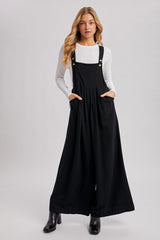 woman wearing a black wide-leg overall dress with pockets and gold button details over a white long sleeve top
