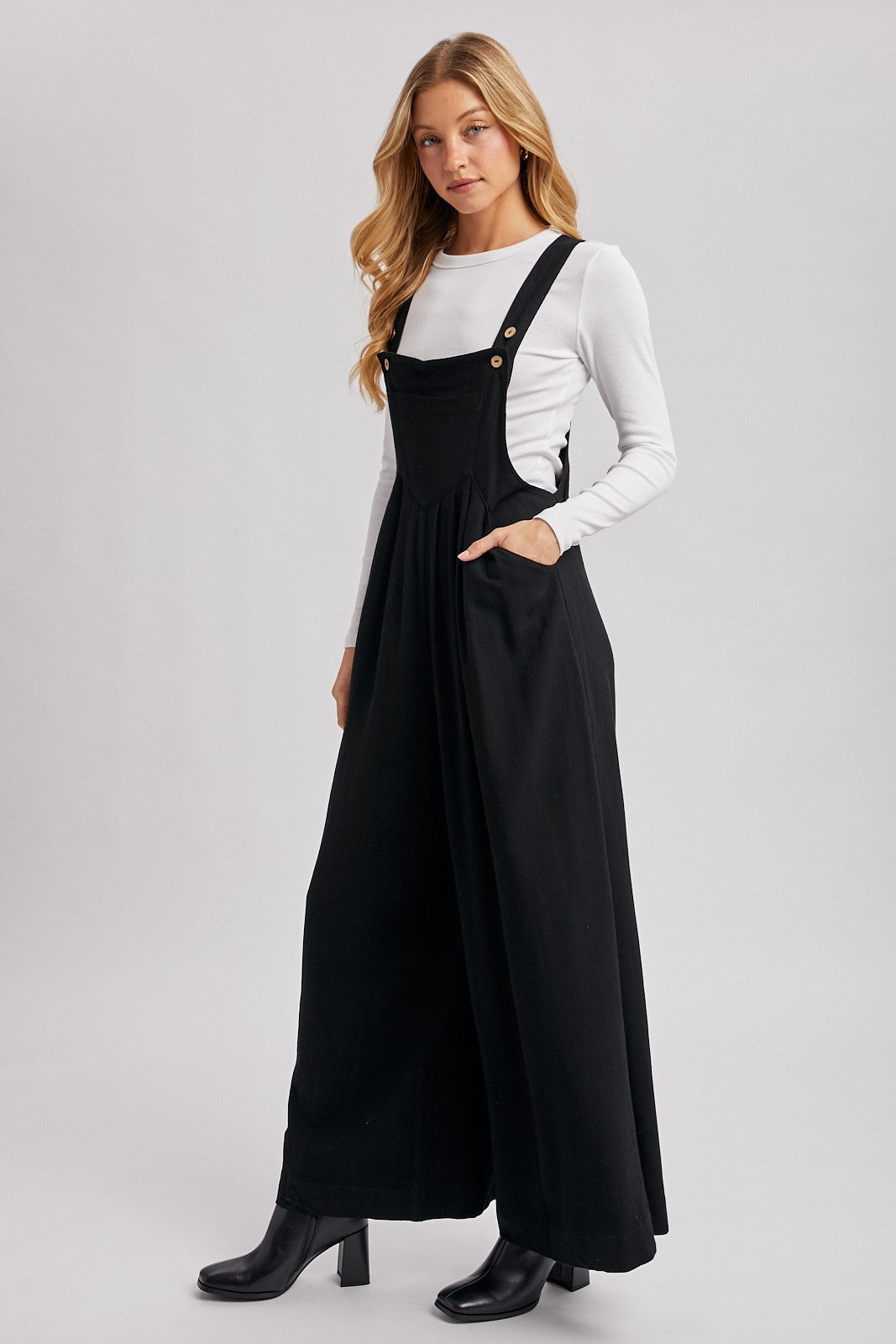 woman wearing a black maxi overall dress with pockets over a white long sleeve top and black heeled boots
