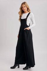 woman wearing a black maxi overall dress with pockets over a white long sleeve top and black heeled boots