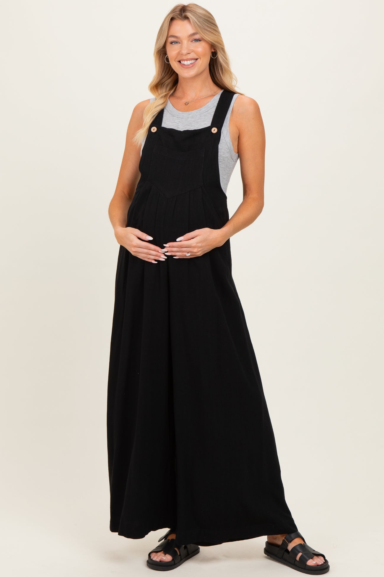 woman wearing a black maternity dungaree dress over a gray top and holding her baby bump