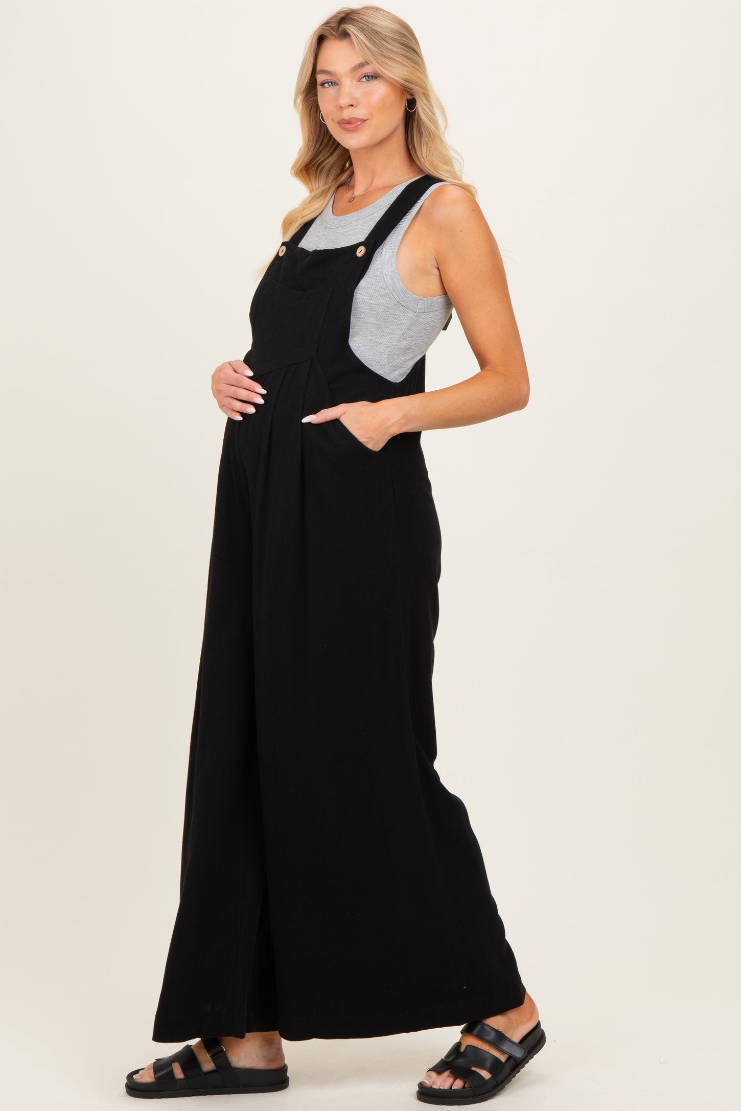 woman wearing a black maternity overall dress with pockets and a gray tank top, showing her baby bump