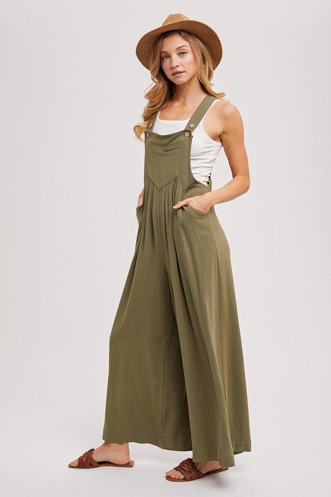 woman wearing an olive green wide-leg jumpsuit with buttons and pockets, paired with a white tank top and a brown hat