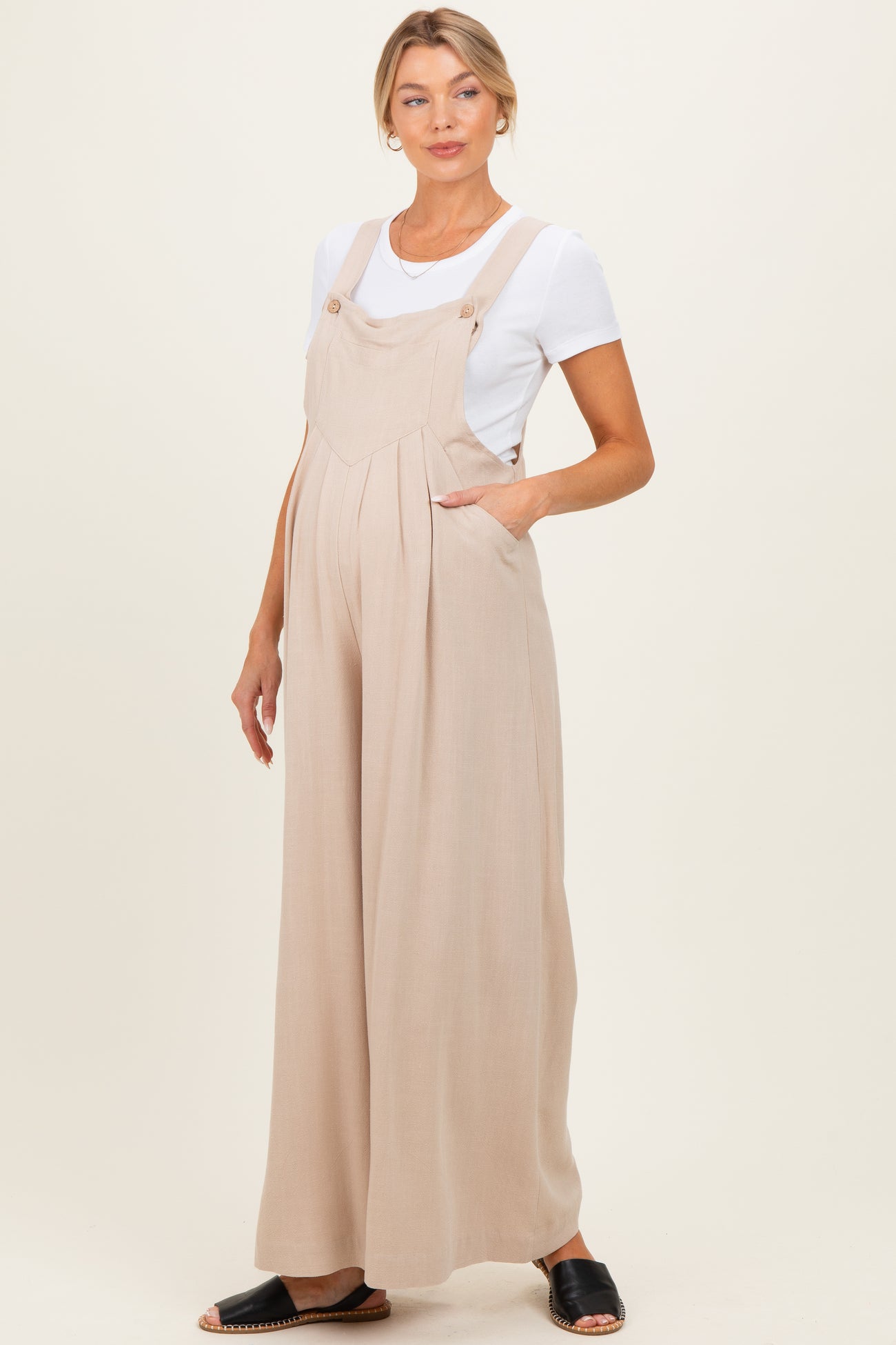 woman wearing a beige maternity jumpsuit with wide legs and side pockets, styled with a white t-shirt underneath and black sandals