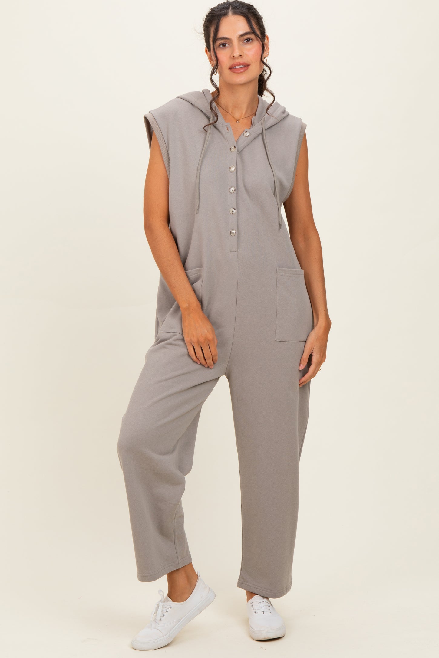 woman wearing a gray sleeveless jumpsuit with a hood and button details, standing against a neutral background