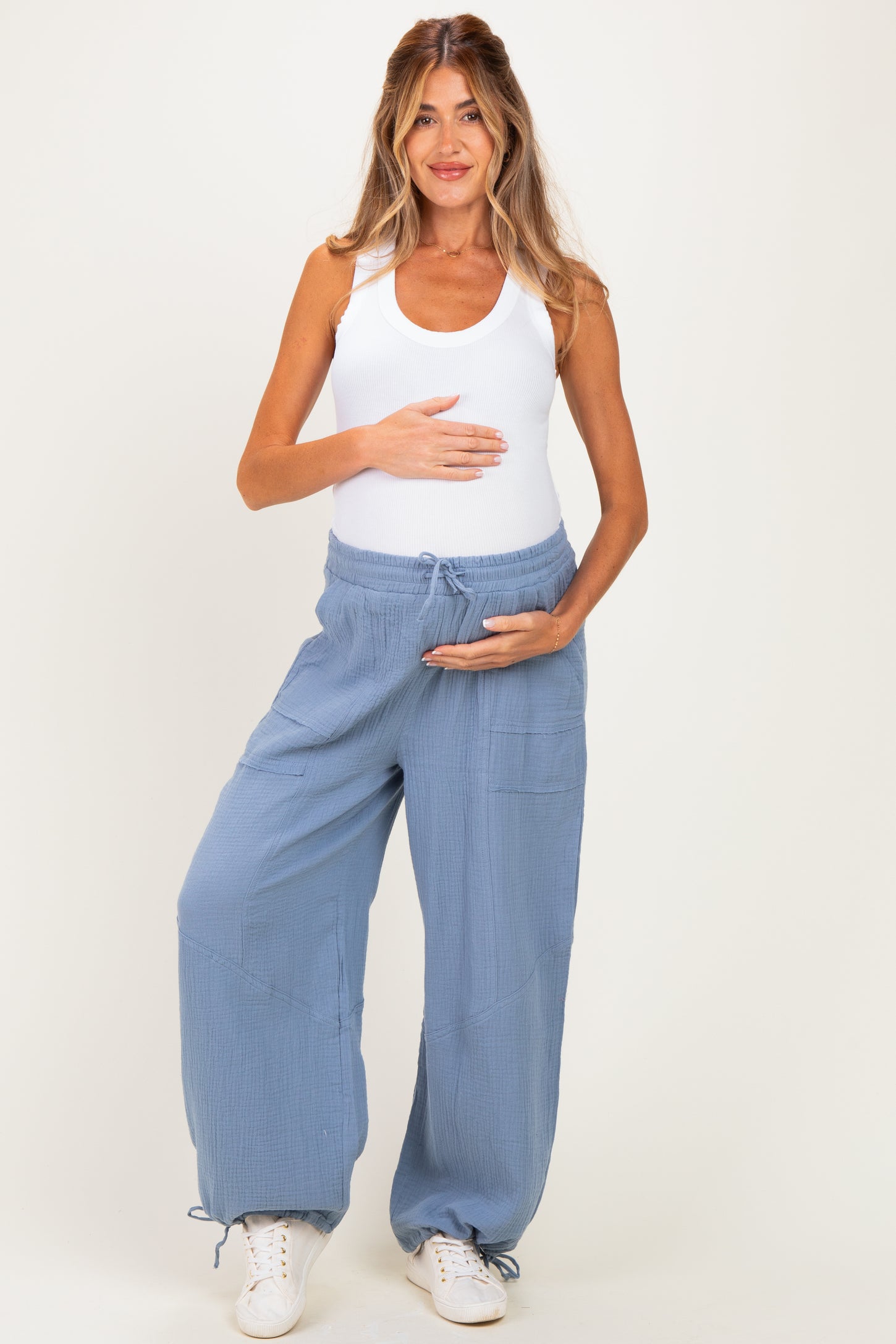 woman wearing a white tank top and light blue maternity pants, holding her baby bump