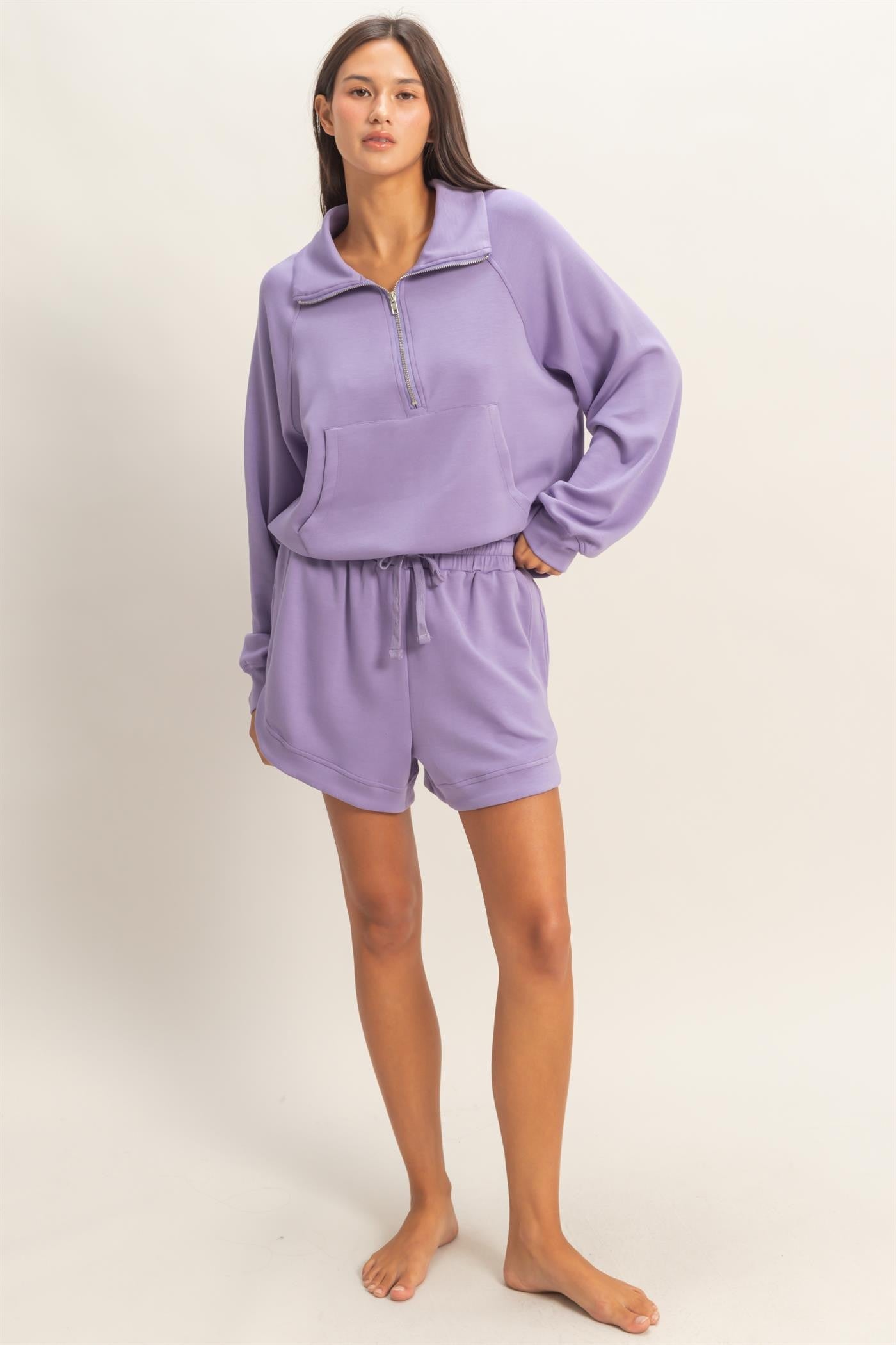 woman wearing a purple zip-up pullover and matching shorts, standing barefoot against a light background