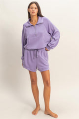 woman wearing a purple zip-up pullover and matching shorts, standing barefoot against a light background
