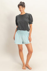 woman wearing a black puff sleeve sweatshirt and light blue smocked shorts, standing barefoot with a neutral background