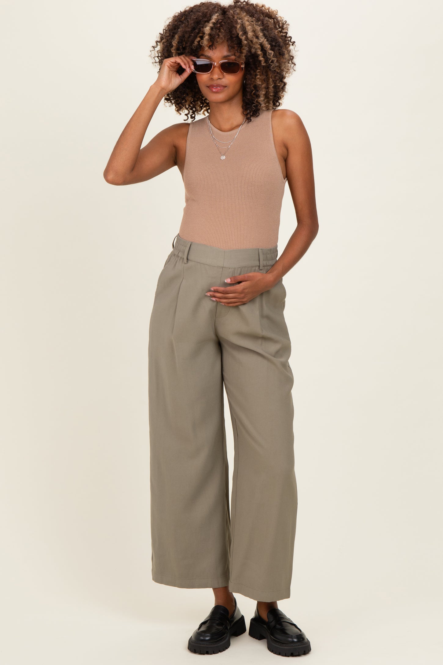 woman wearing a beige ribbed tank top and olive green wide-leg maternity pants, holding her baby bump