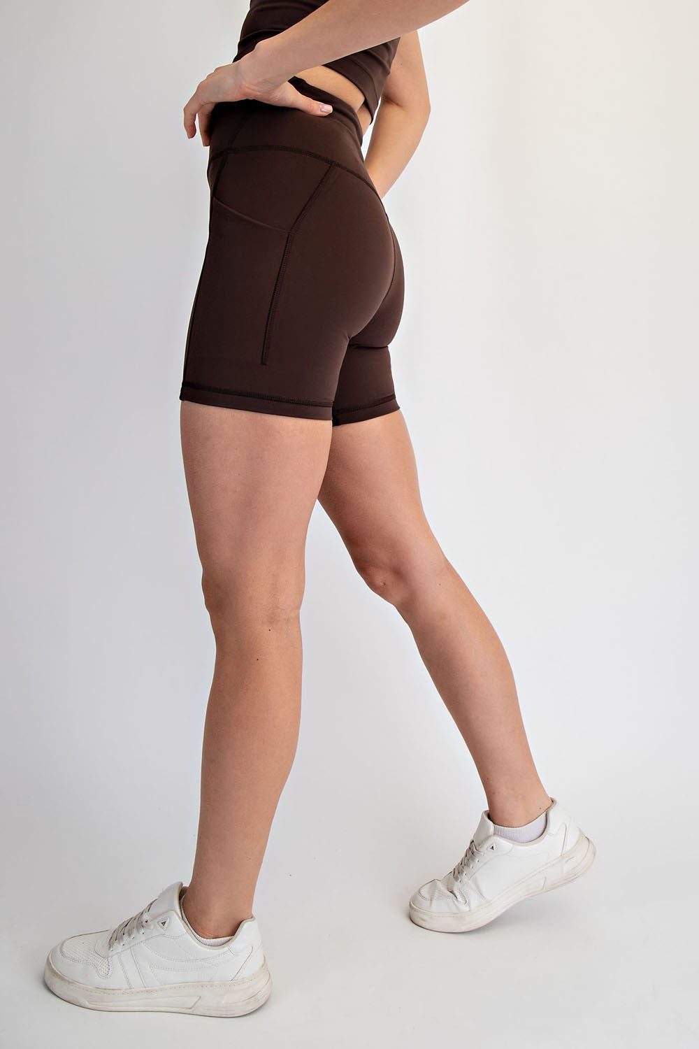 woman wearing brown high-waisted workout shorts and white sneakers standing in profile with a plain background