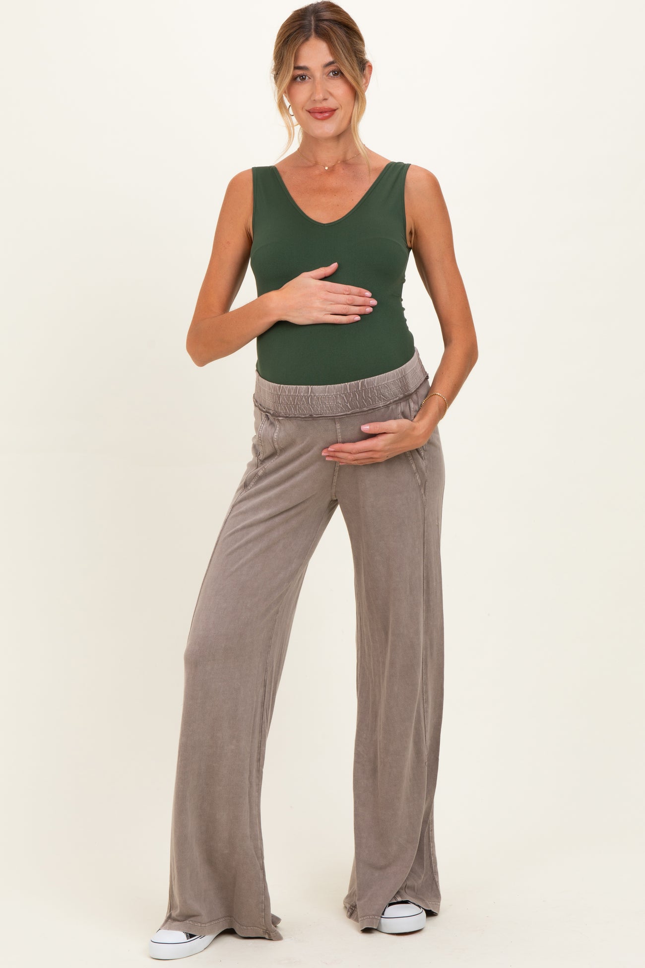 woman wearing a green sleeveless maternity top and beige wide-leg maternity pants, holding her baby bump