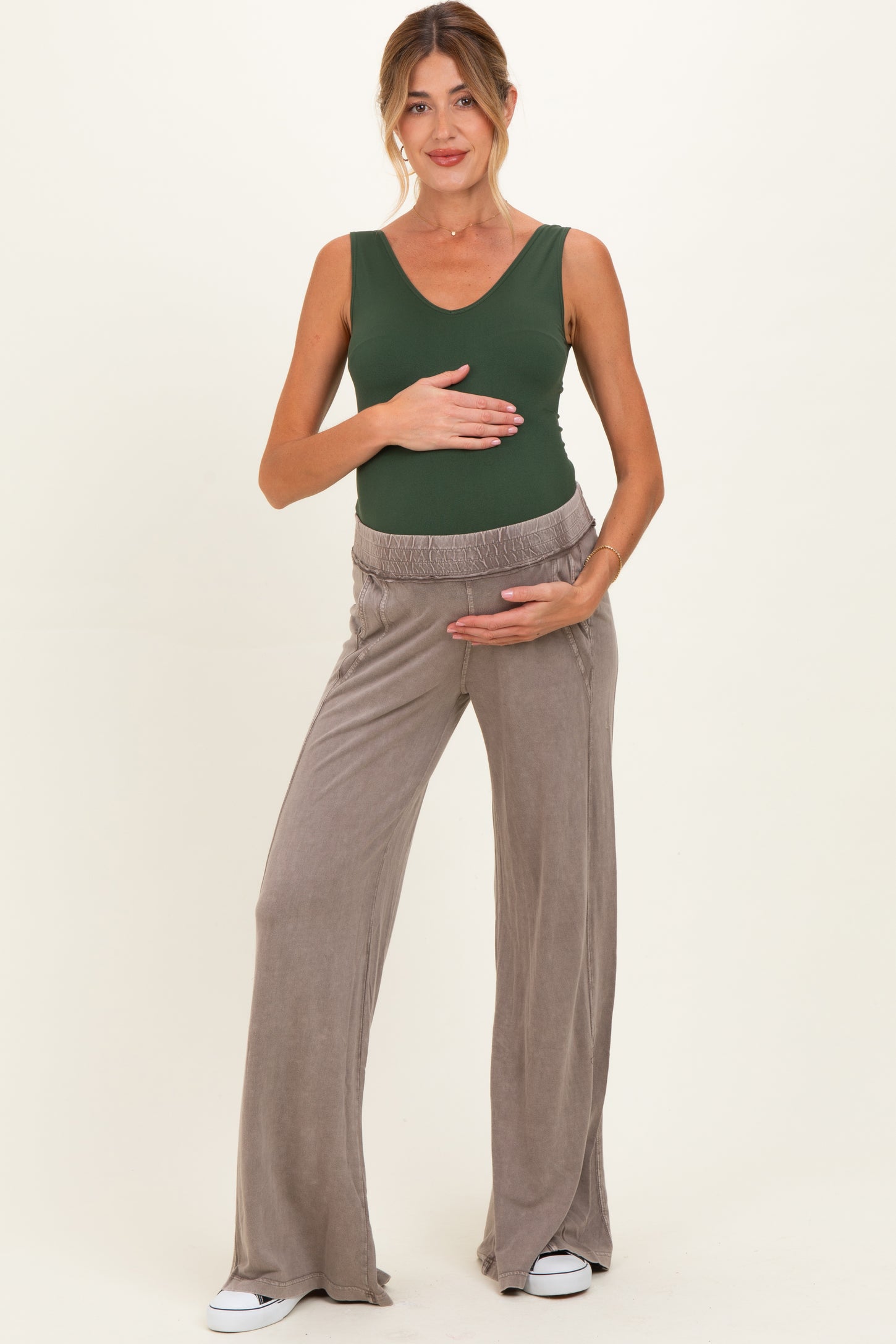 woman wearing a green sleeveless maternity top and beige wide-leg maternity pants, holding her baby bump