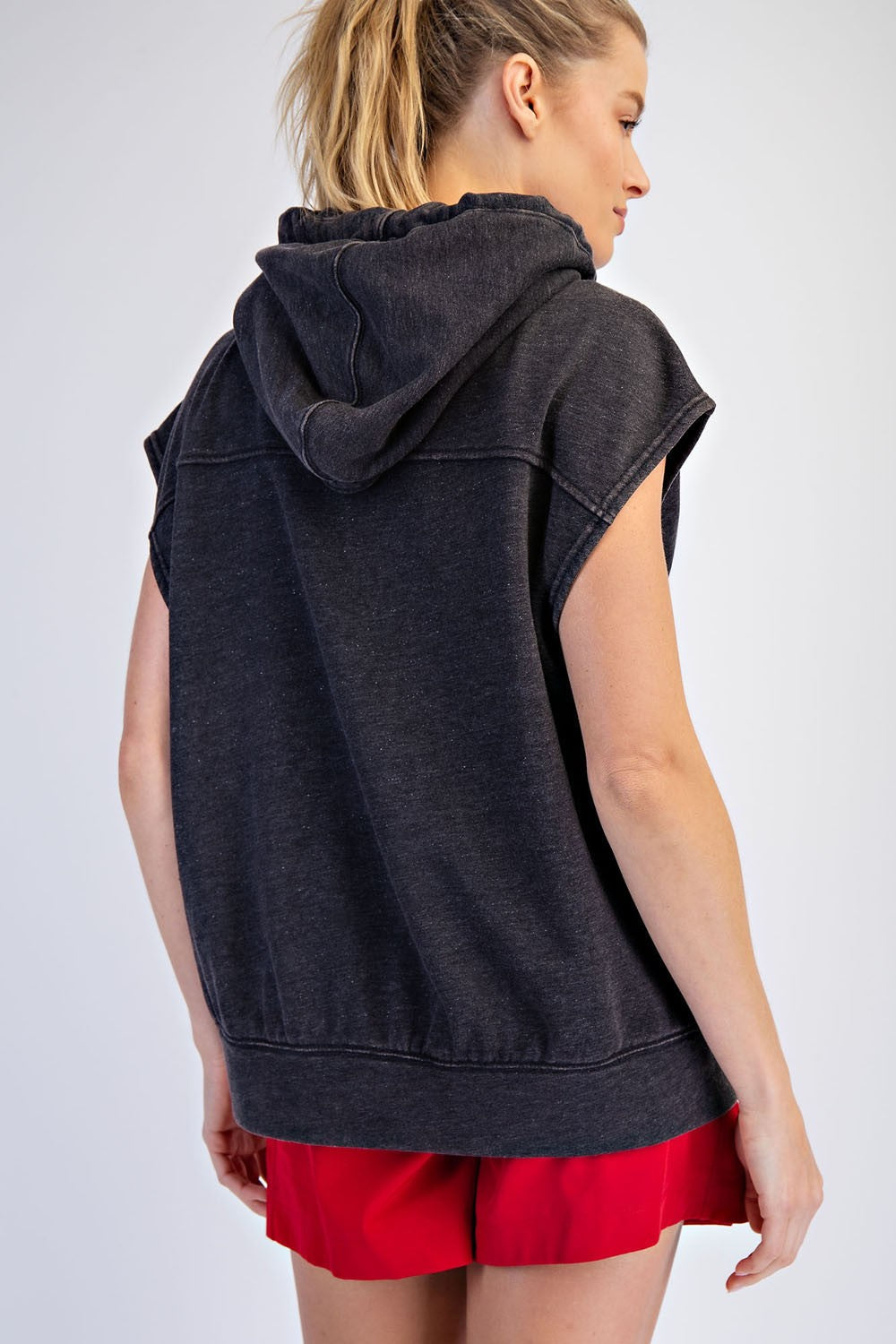 woman wearing a dark gray sleeveless hoodie with a hood and red shorts, viewed from the back