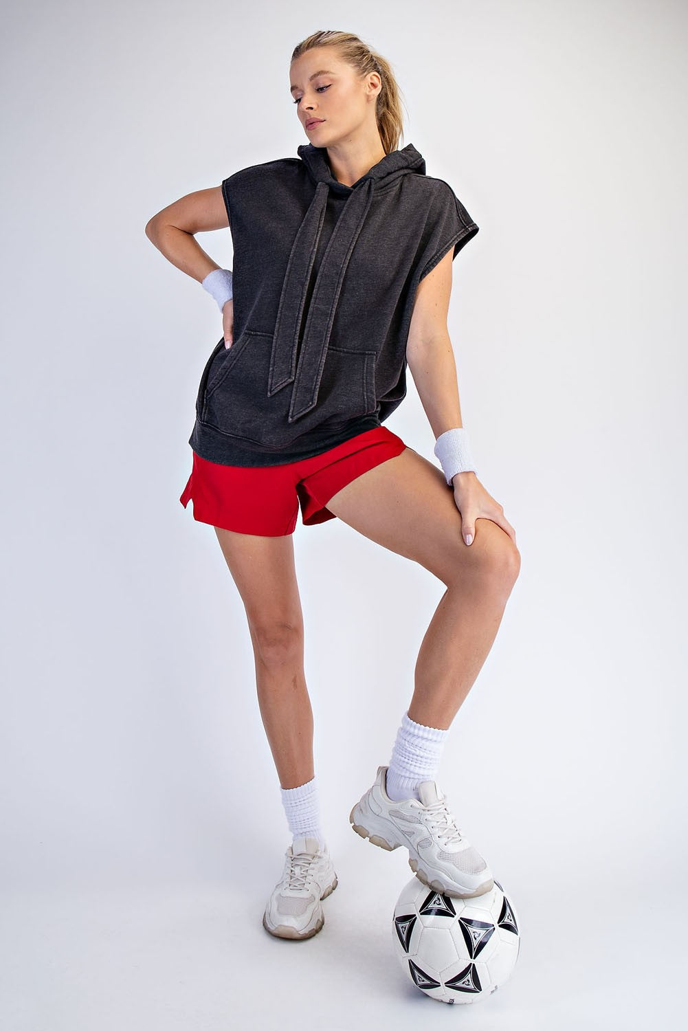 woman wearing a dark gray sleeveless hoodie and red athletic shorts, posing with a soccer ball