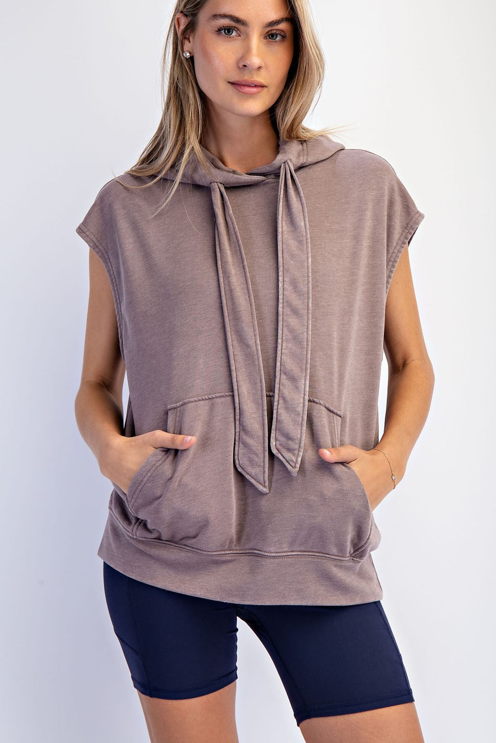 woman wearing a taupe sleeveless hoodie with a front pocket and tie detail, paired with navy bike shorts