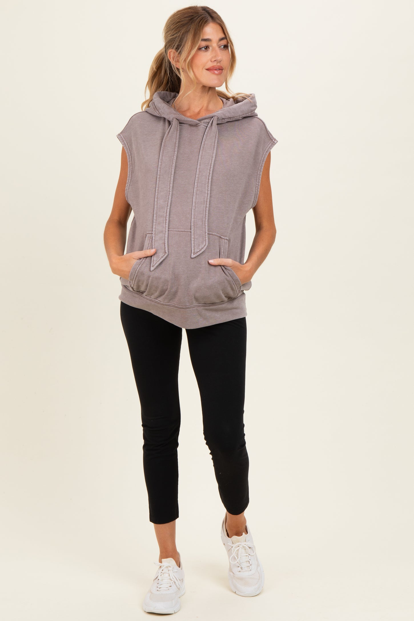 woman wearing a gray sleeveless maternity hoodie with pockets and black leggings, showing her baby bump