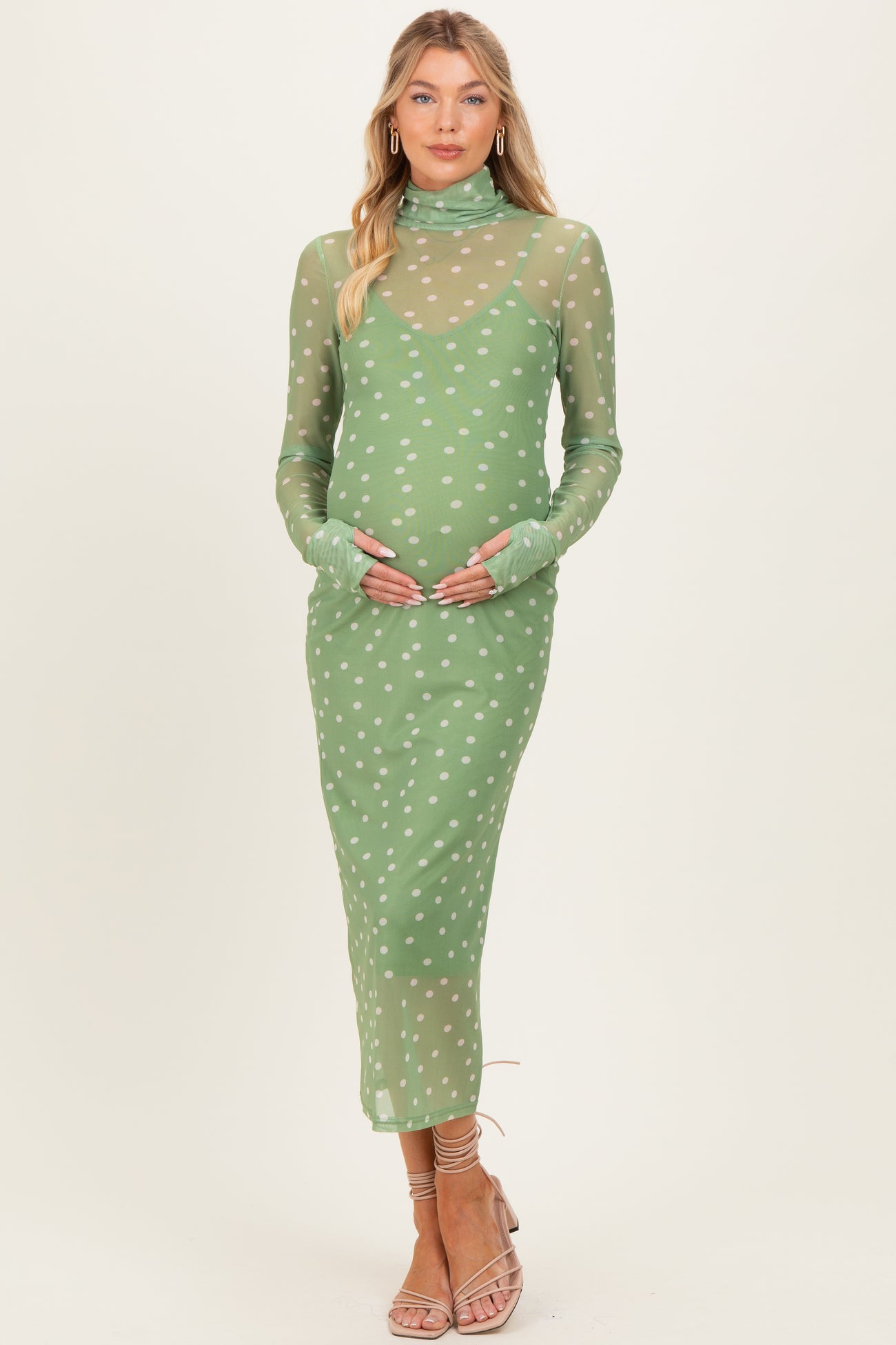 woman wearing a green sheer maternity dress with white polka dots and long sleeves, gently cradling her baby bump