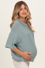 woman wearing a light green oversized maternity sweater, gently holding her baby bump