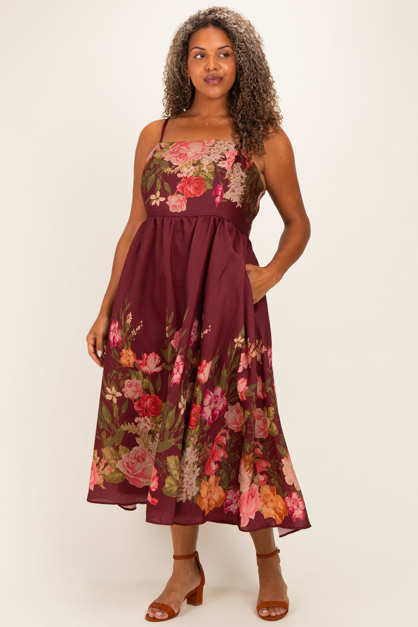 woman wearing a burgundy floral plus size midi dress with a fitted bodice and pockets, styled with brown heeled sandals