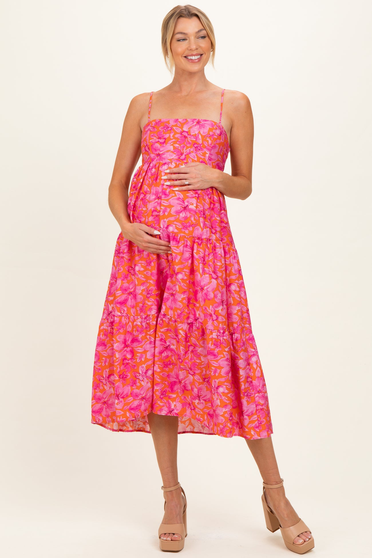 woman wearing a vibrant pink floral maternity dress with adjustable straps and holding her baby bump