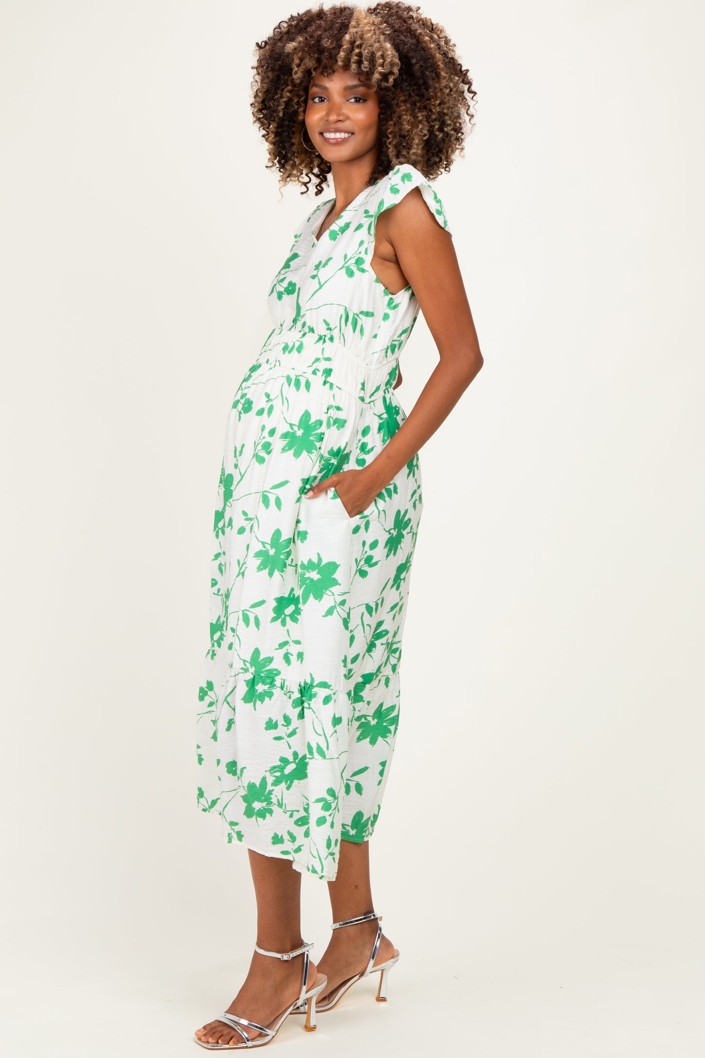 woman wearing a green and white floral maternity dress with adjustable straps and pockets, showcasing her baby bump