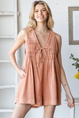 woman wearing a rust-colored sleeveless romper with pleats and a tie front, posing with a smile and hands in pockets