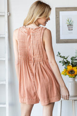 woman wearing a rust-colored sleeveless romper with pleated back detail, standing indoors near a flower vase