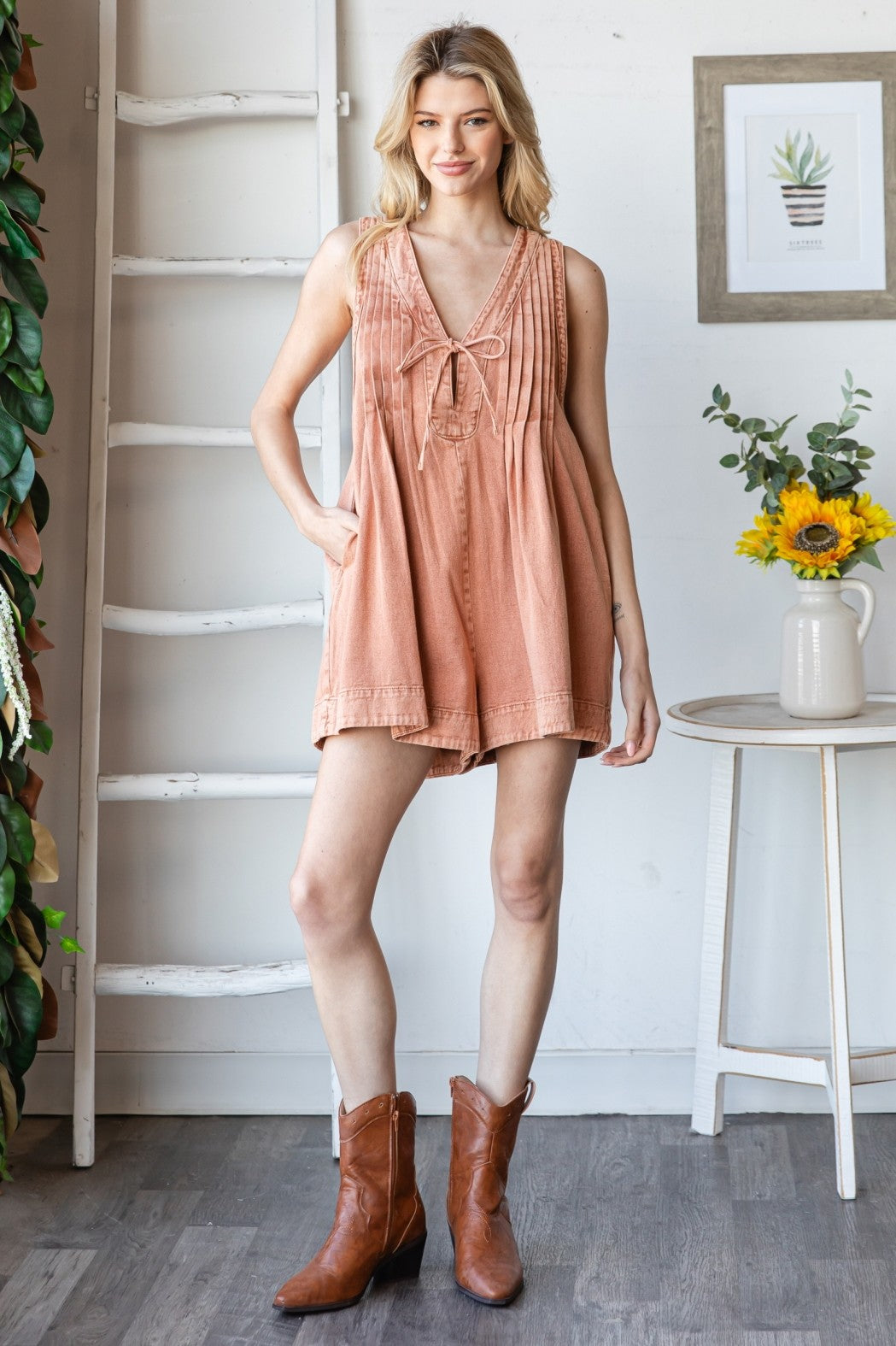 woman wearing a rust-colored sleeveless romper with a tie detail and pockets, paired with brown cowboy boots