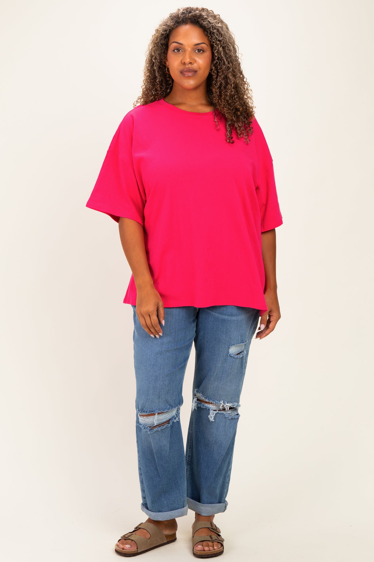 woman wearing a bright pink oversized t-shirt and distressed blue jeans, standing against a neutral background