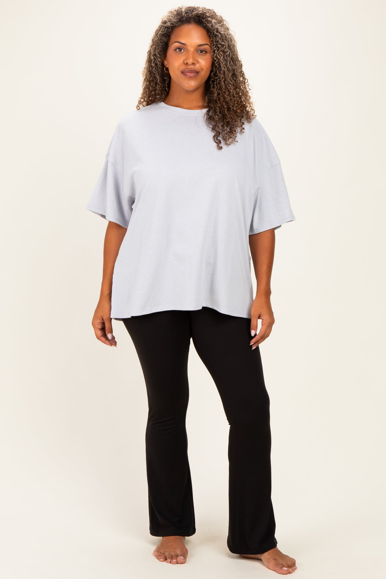 woman wearing a light gray oversized t-shirt and black flared pants, standing barefoot against a neutral background