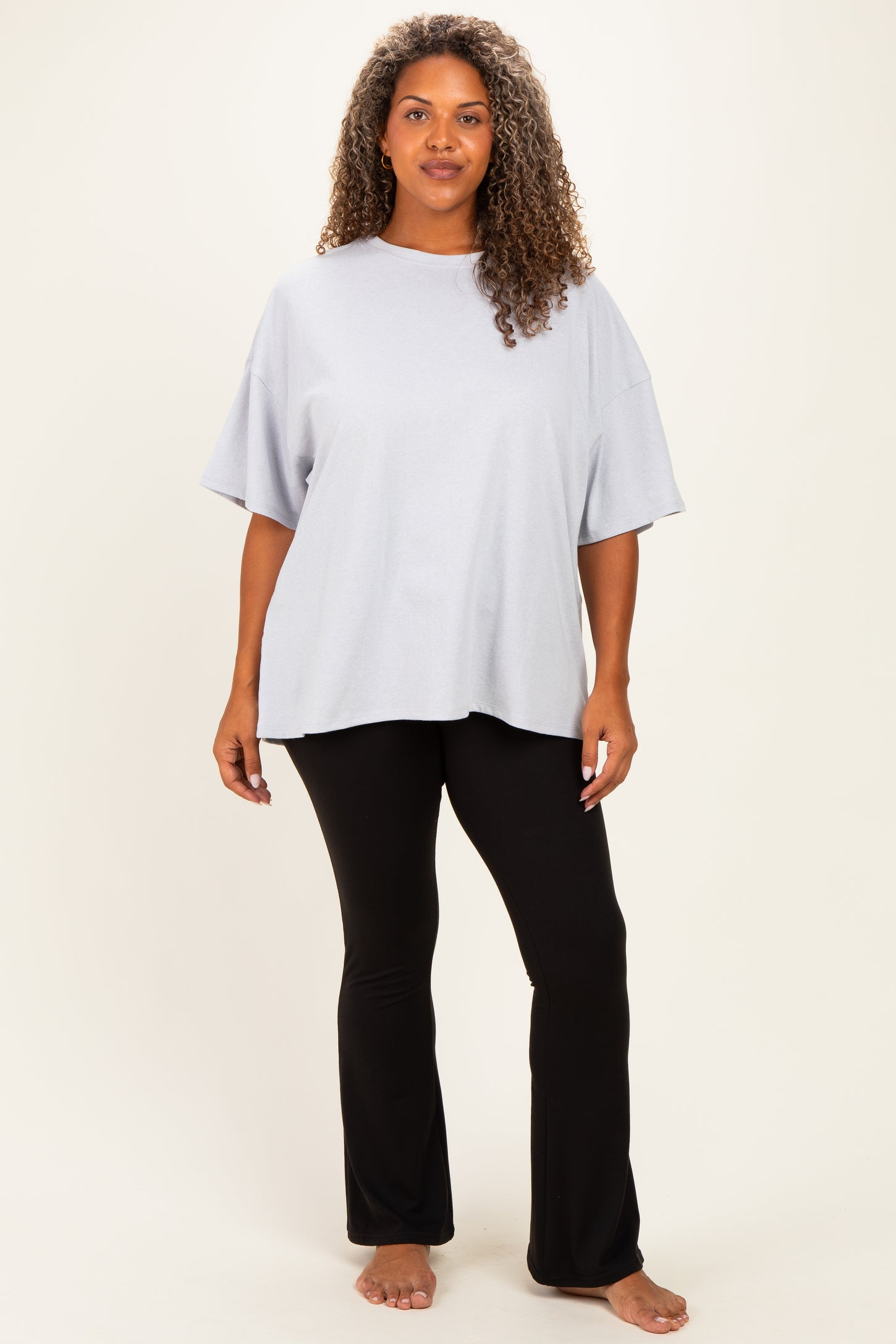 woman wearing a light gray oversized t-shirt and black flared pants, standing barefoot against a neutral background