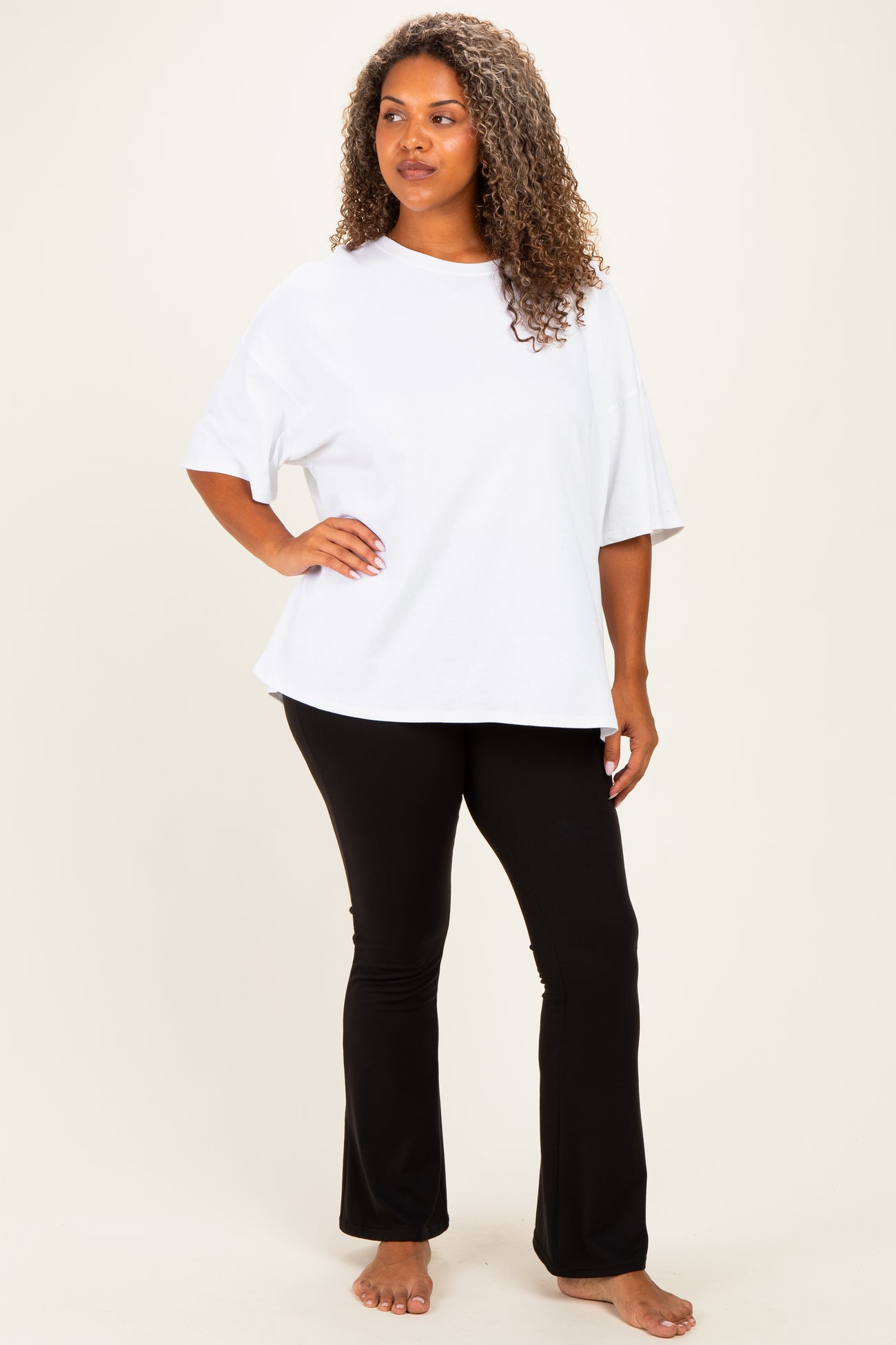 woman wearing a white oversized t-shirt and black flared pants, standing barefoot against a light background