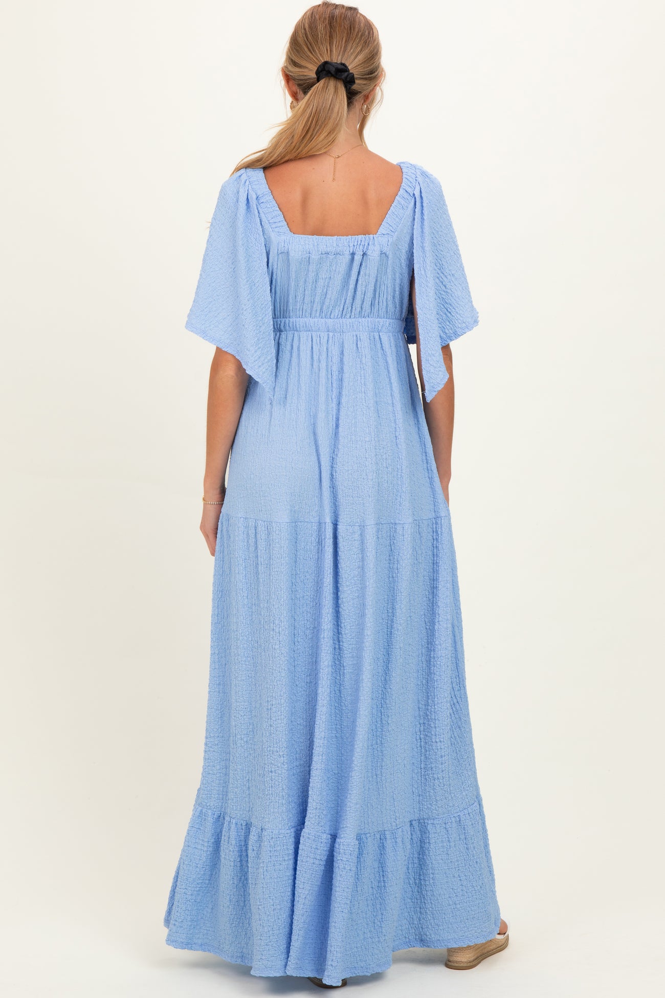 woman wearing a back view of a light blue maternity maxi dress with flutter sleeves and tiered skirt