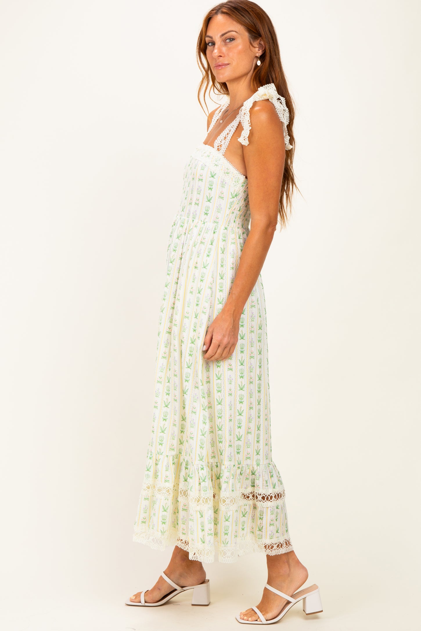 woman wearing a white maxi dress with green floral prints and lace details, styled with strappy sandals