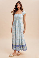 woman wearing a light blue floral maxi dress with a smocked bodice and ruffled hem, standing on a neutral background