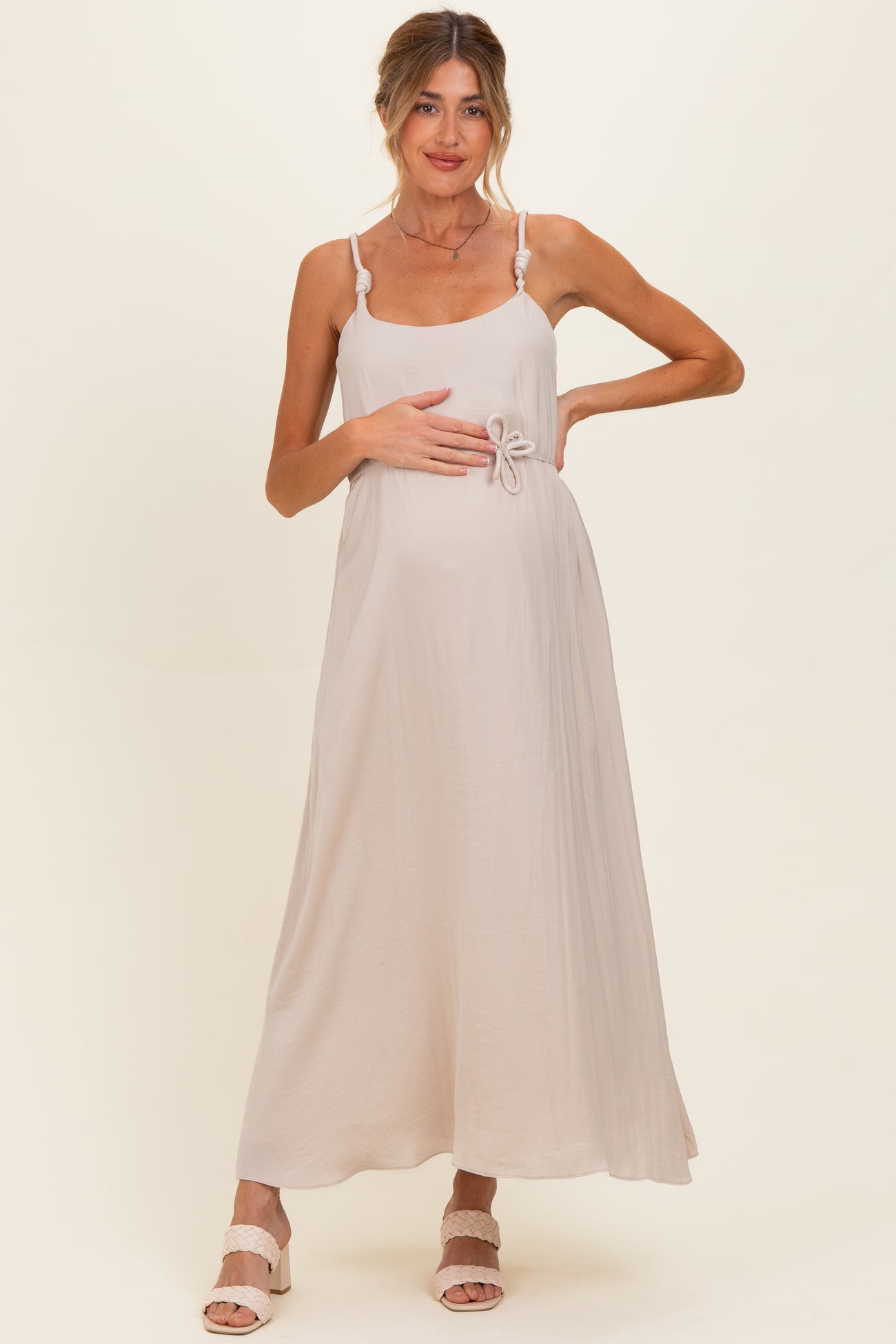 woman wearing a light beige maternity maxi dress with a tie detail at the waist and holding her baby bump