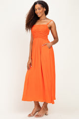 woman wearing a bright orange maxi dress with smocked bodice and adjustable straps, standing with one hand in pocket