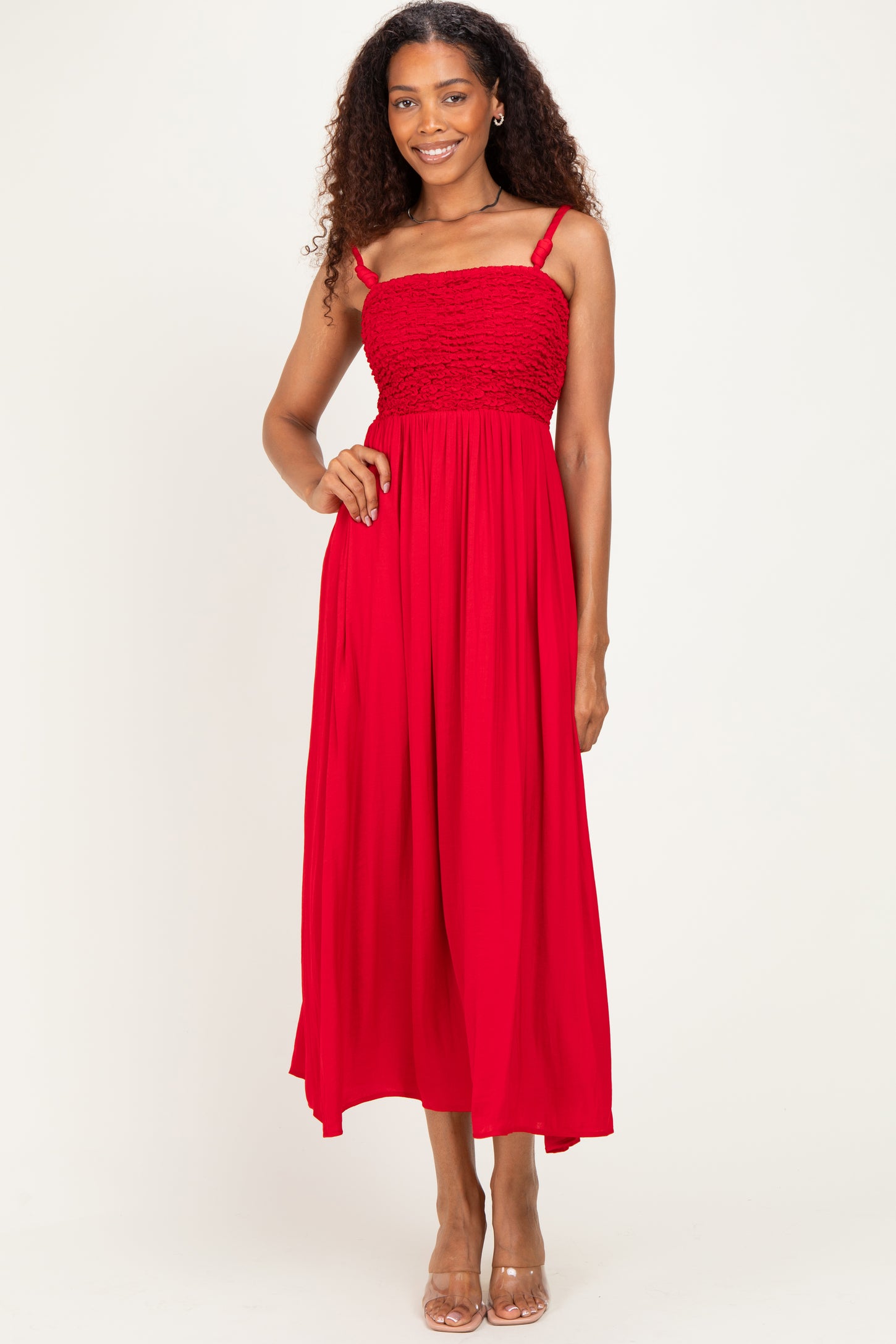 woman wearing a red maxi dress with smocked bodice and thin straps, posing with one hand on her hip