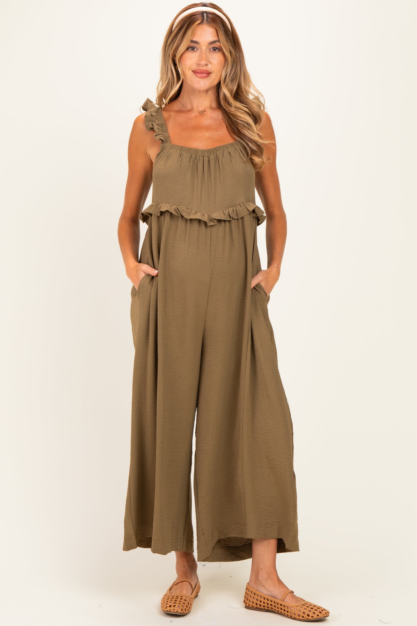 woman wearing a khaki green maternity jumpsuit with ruffle details and pockets, showing her baby bump