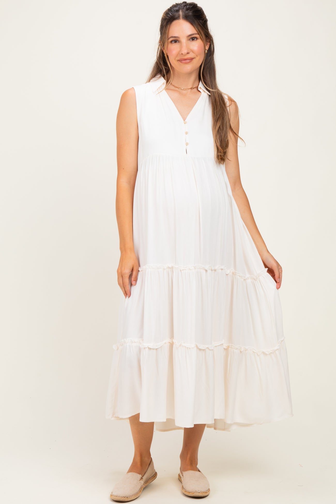 woman wearing a white sleeveless maternity dress with a button closure and tiered ruffle hem, showing her baby bump