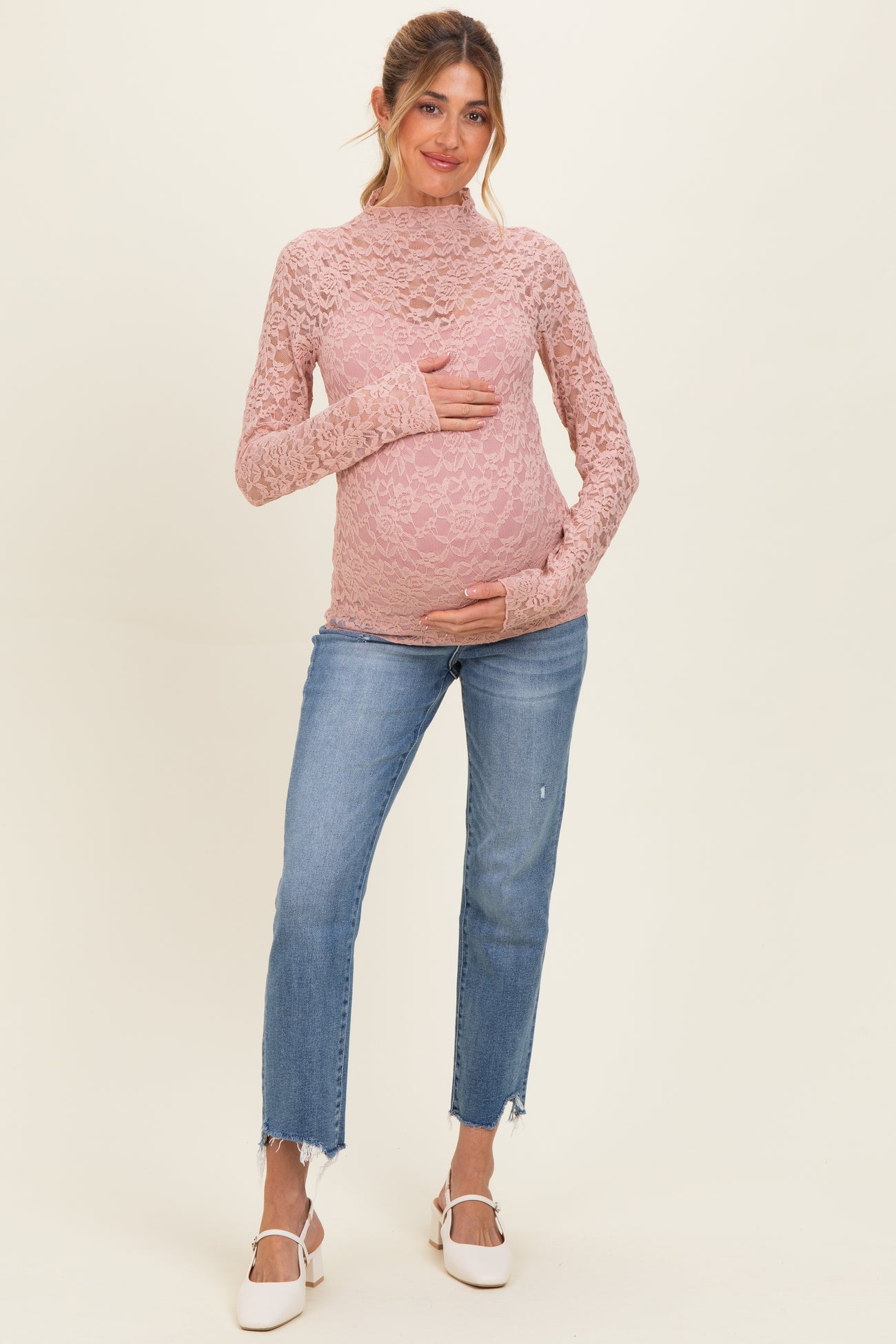 woman wearing a pink lace long sleeve maternity top and holding her baby bump with distressed blue jeans